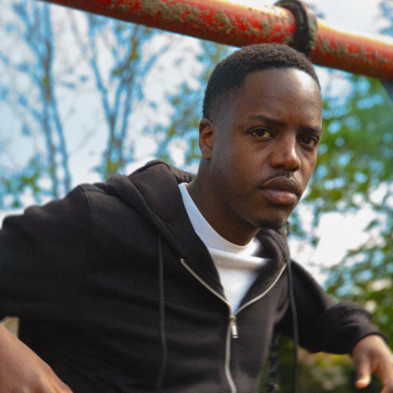 Performer stands in a park during the day, wearing a black hoodie