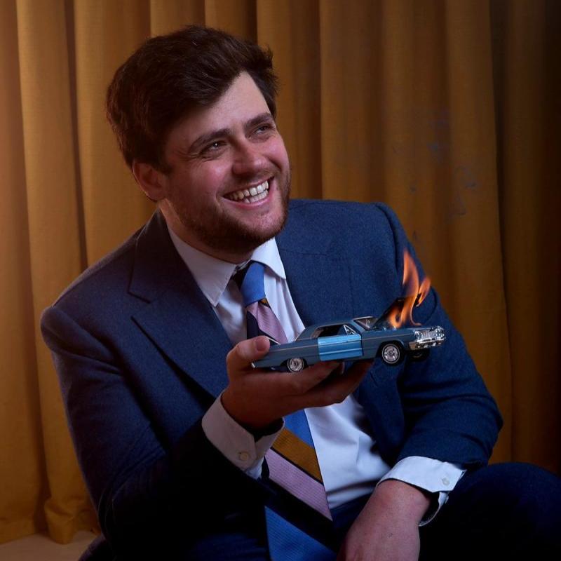 Bróccán Tyzack-Carlin, a man with brown hair and stubble, is wearing a blue suit and white shirt with a colourful tie. He is smiling looking to the side and holding a miniature car with the hood on fire, over a brown curtain background.