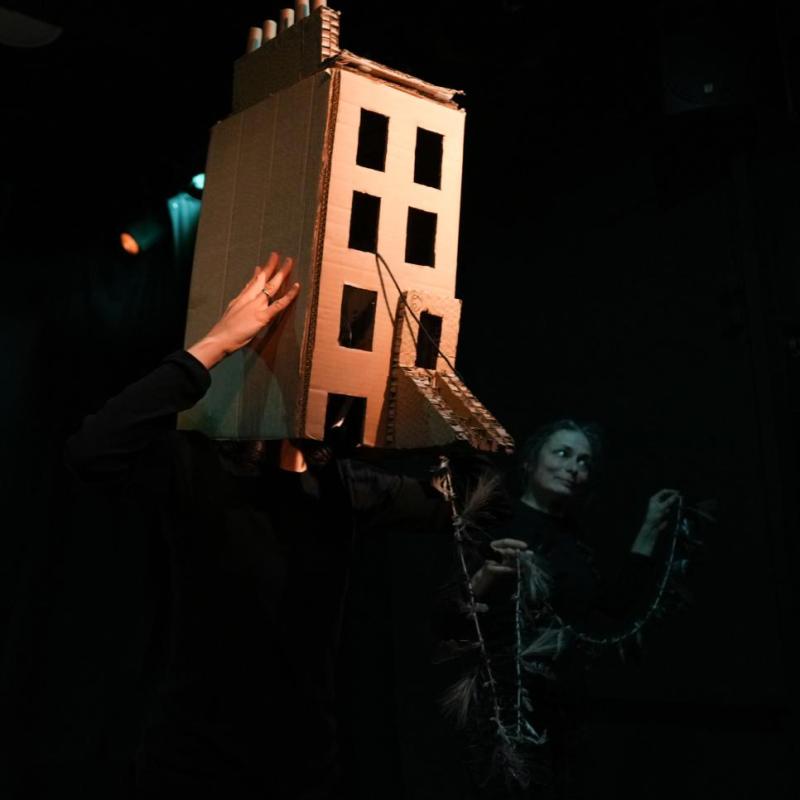 Performers in the dark hold up a brightly lit cardboard townhouse