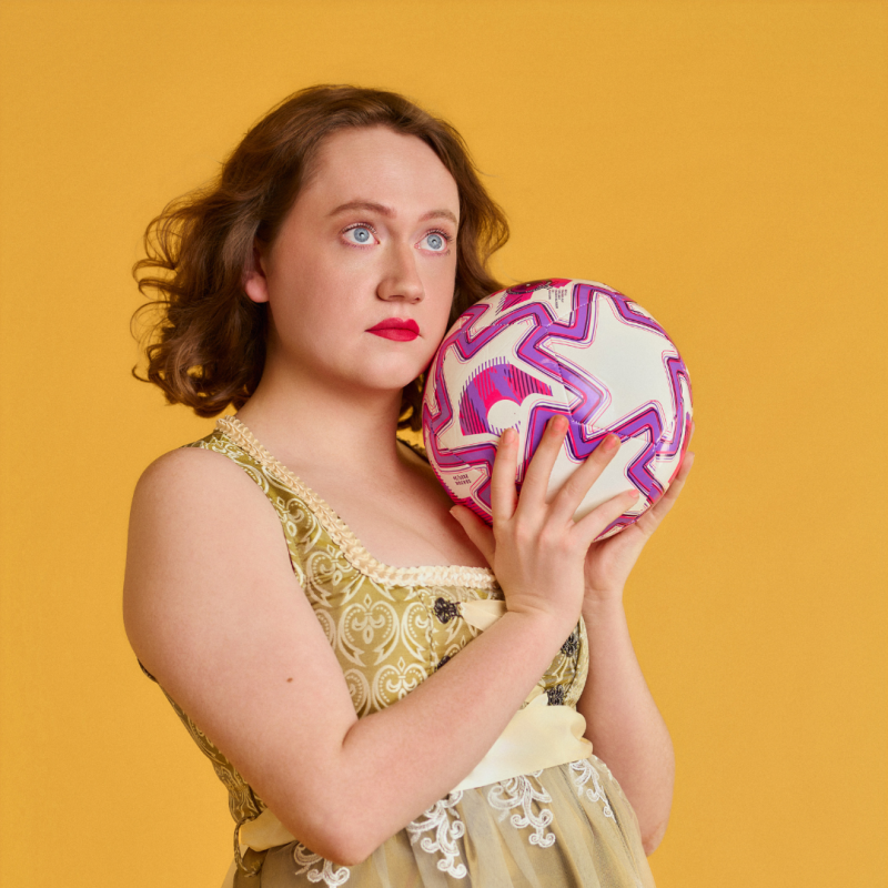 Dee stands in front of a yellow back drop. They are holding white, pink and purple football and gazing into the distance.