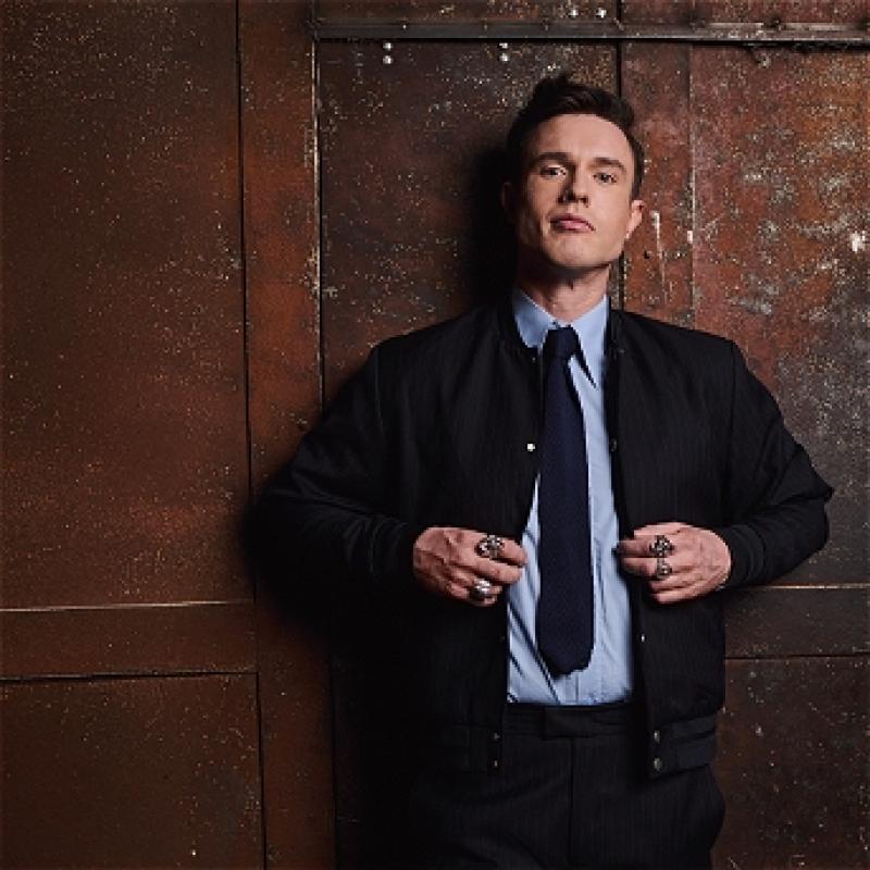 Ed Gamble leans against a wooden wall. He is smartly dressed in a blue shirt and black tie, and looks confidently into the camera.