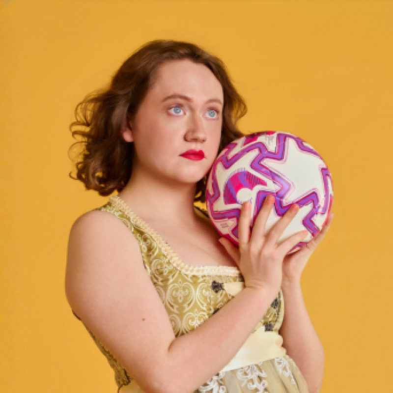 Dee stands in front of a yellow back drop. They are holding white, pink and purple football and gazing into the distance.