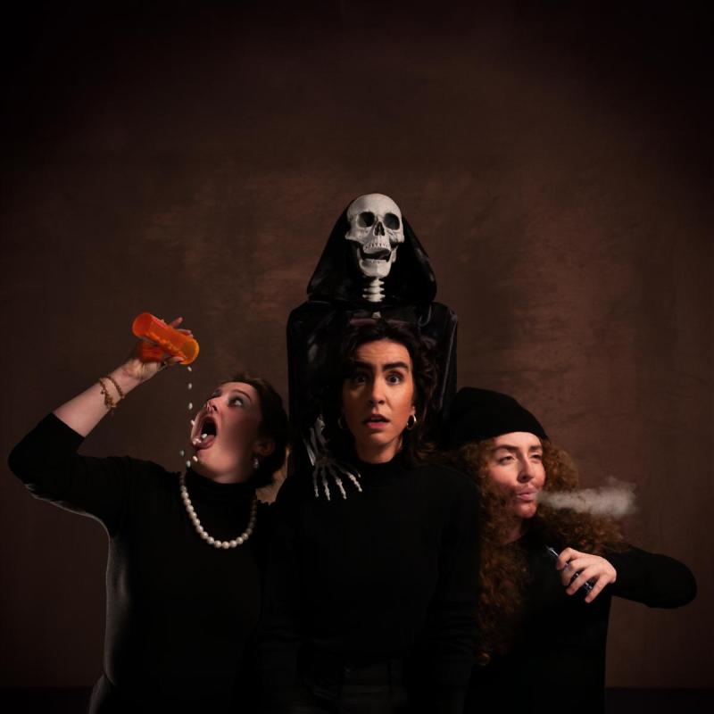 Three performers stand before a skeleton in a robe: the one in the center looks straight ahead, the one on their left smokes, and the one on their right pours a bottle of pills into their mouth, in front of a brown background.