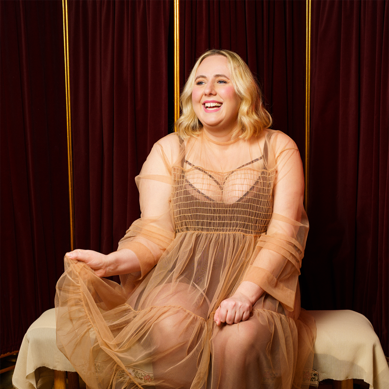 Helen sits on a bench, dressed in a sheer flowy dress. They smile happily. 