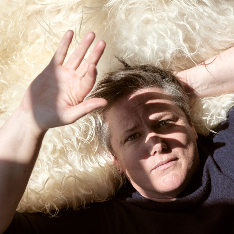 Hannah Gadsby lies on a bed of white fur. They look up at the camera and block the sunlight with their hand.