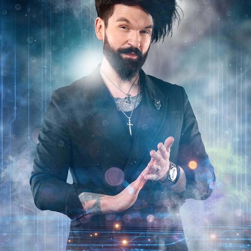 Colin Cloud, wearing a black suit jacket with chain necklaces, rubs his fingertips of one hand onto the palm of his other. A ball of light emenates from his palm. He is surrounded by smoke. 
