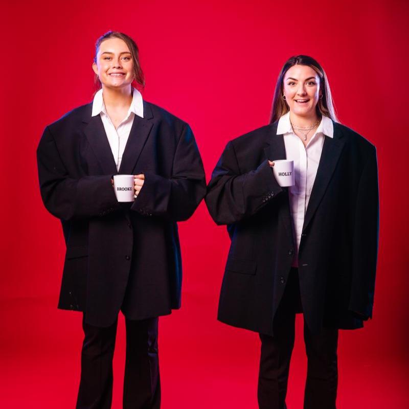 Holly and Brooke stand against a red background, wearing oversized suits