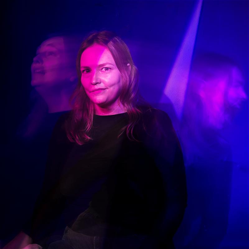 Kate is looking into the camera. A beam of light illuminates half of her face. 