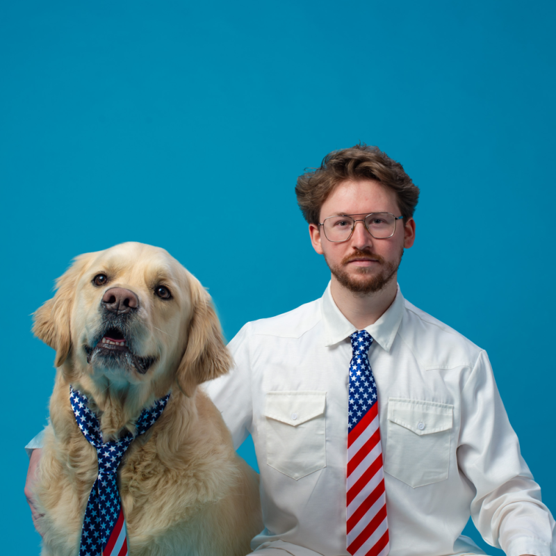 A man and his golden retrierver sit in front of a bright blue background. He wears a shirt and an American flag tie. His dog also wears an American flag tie.  