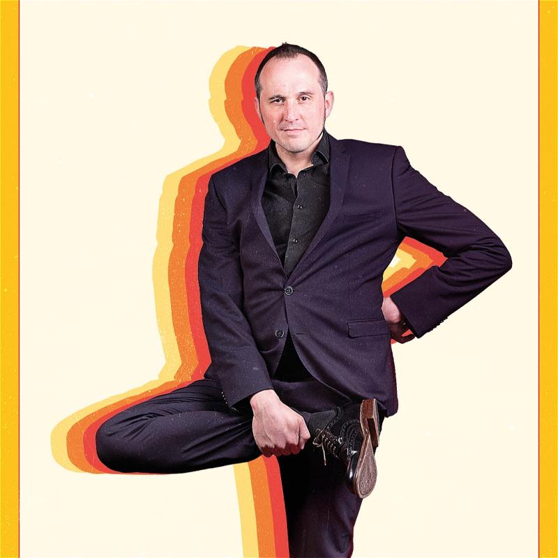 Phil looks into the camera whilst holding up their right foot. Behind them are colourful silhouettes. 