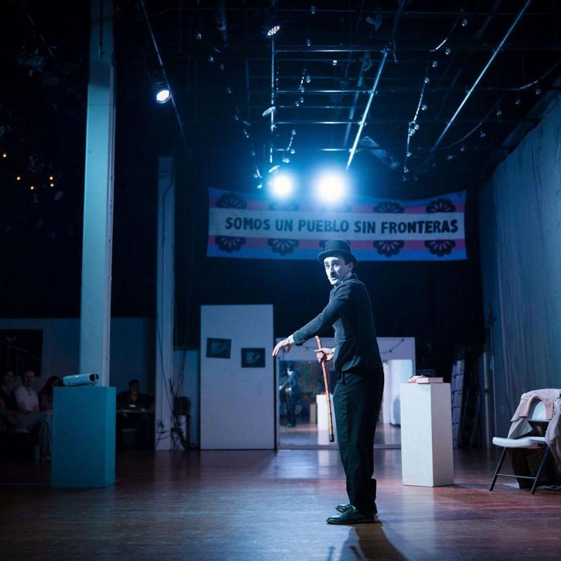A wide shot of a young man dressed as Charlie Chaplin. He has a white face, black eyebrows and toothbrush moustache, and wears a white shirt, black jacket and bowler hat and holds a bamboo cane in one hand. He stands on stage.