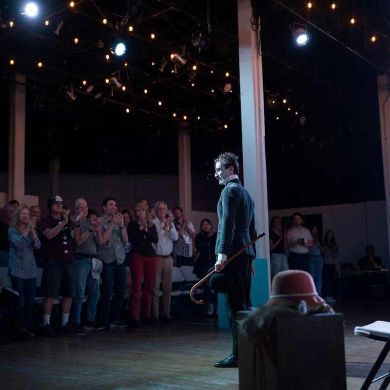 A wide shot of a young man dressed as Charlie Chaplin. He has a white face, black eyebrows and toothbrush moustache, and wears a white shirt, black jacket and bowler hat and holds a bamboo cane in one hand. He stands on stage receiving a standing ovation..