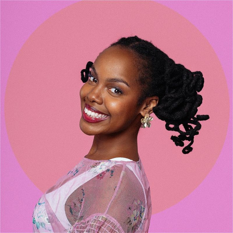Sharon is standing side on, smiling at the camera against a pink background
