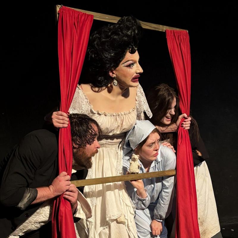 The performers huddle together around a window frame prop in front of a black background