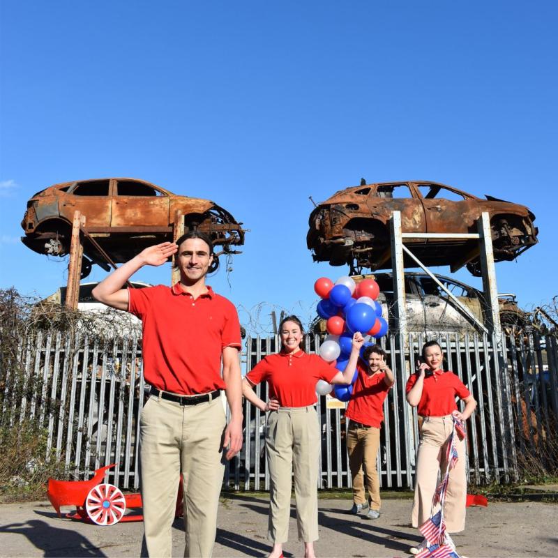 Four performers wearing red polos and khaki pants stand in front of a car junkyard