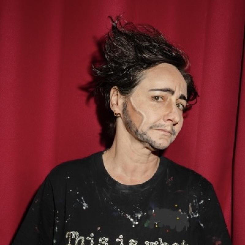 The performer stand before a red velvet curtain wearing a paint-splattered shirt that says "this is what a feminist looks like." in rhinestones.