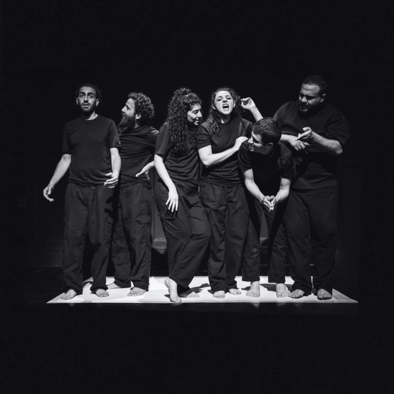A group of performers wearing black stand on a white stage, surrounded by darkness. 