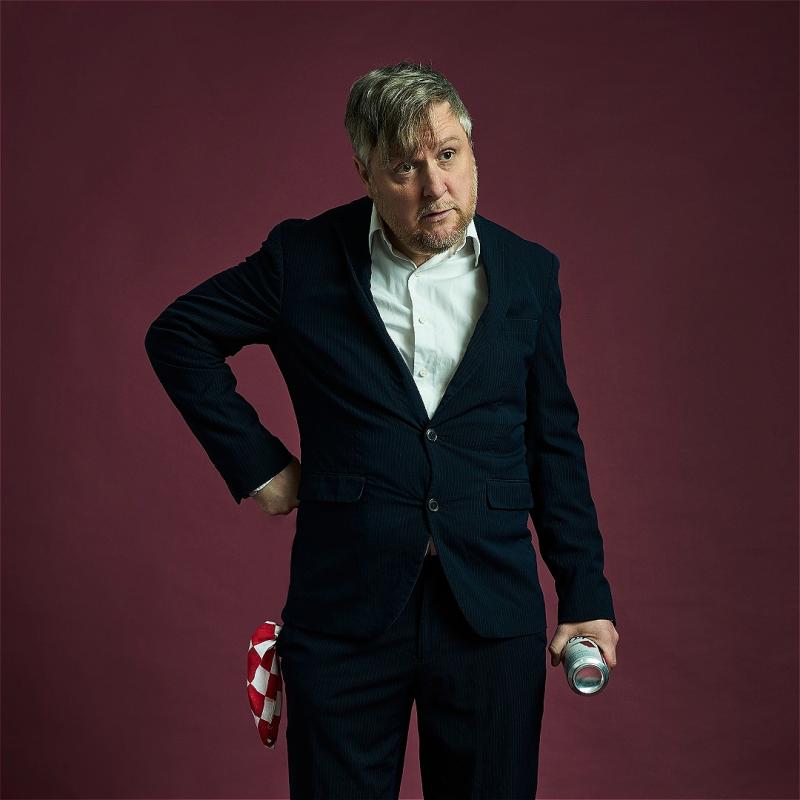Tim Key stands in a suit with his hand on his hip against a purple backdrop