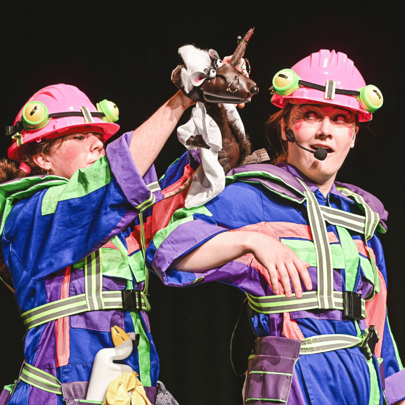 2 performers dressed as frogs in brightly coloured boilers suits. A puppet of a rat scientist travels on the shoulder of one of the performers, waving it's arm as if it is giving directions