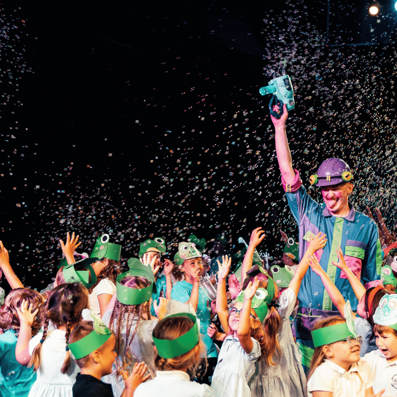 A performer dressed as a frog in a brightly coloured boiler suit holds a bubble gun in the air. They are surrounded by dancing children in frog headbands