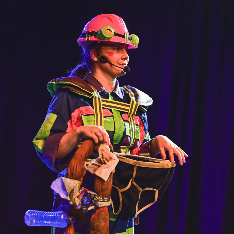 A performer dressed as a frog in a brightly coloured boiler suit is holding a big pair of dark blue Y-Fronts and looking disapprovingly at the audience