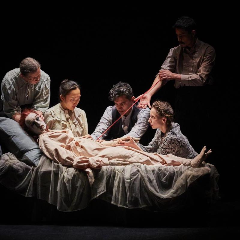 Photo of people surrounding a puppet lying down on a bed