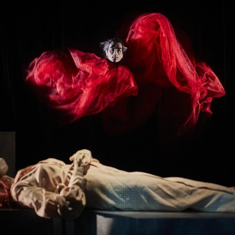 This image shows a female puppet lying flat with a figure with a red cape above her