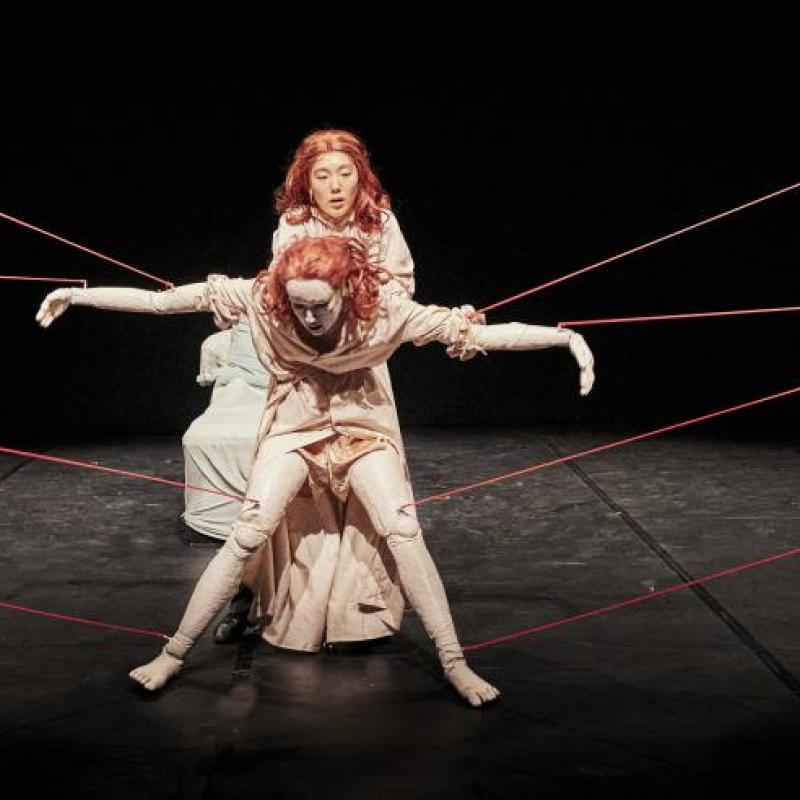Photo of a girl holding a puppet in the centre, with puppeteers pulling the ropes connected to the doll
