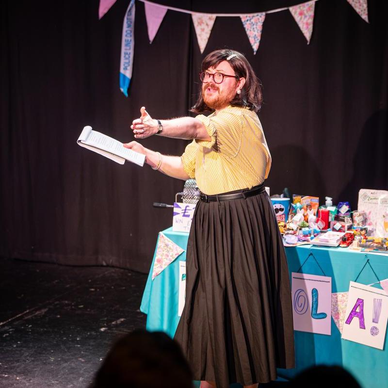 A white person with a brown bob, beard and purple glasses is wearing a yellow blouse and black skirt. They are stood in front of a well-stocked tombola table and are gesturing out into the audience.