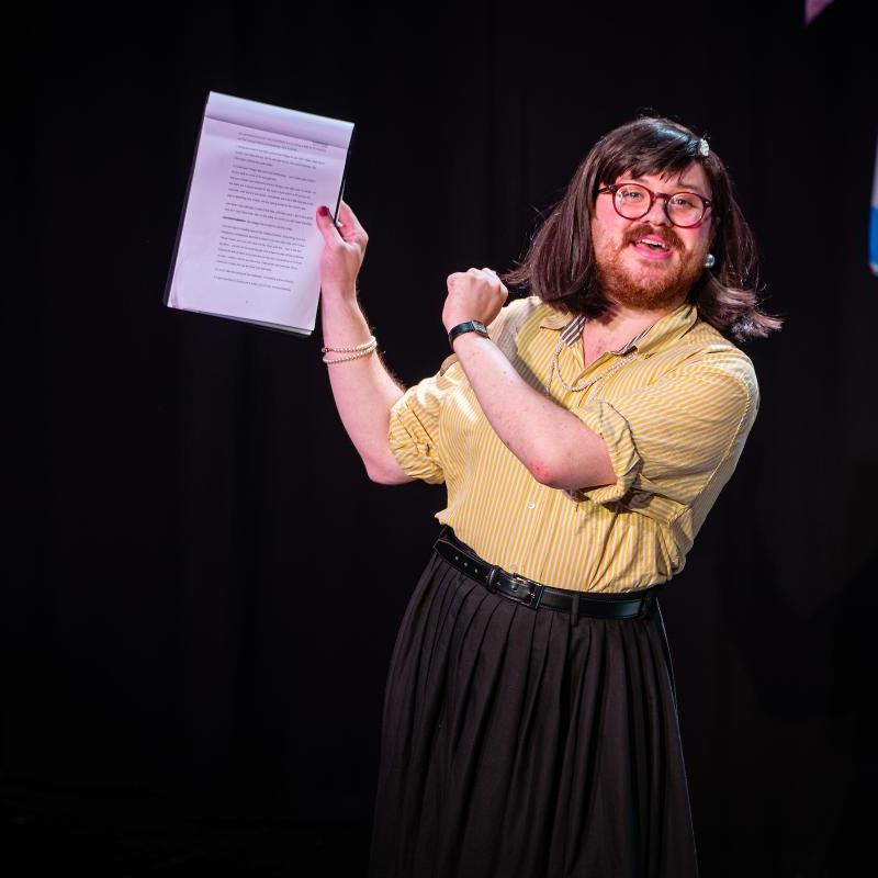 A white person with a brown bob, beard and purple glasses is wearing a yellow blouse and black skirt and gesticulating wildly with a clipboard