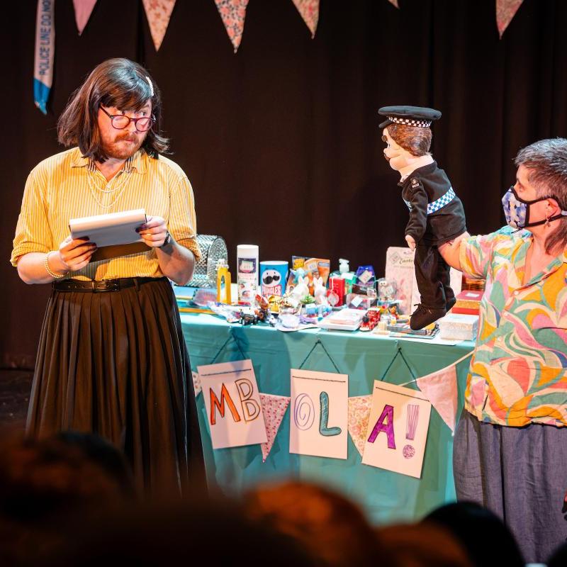 Two people are stood in front of a tombola table on has a brown bob and is wearing a yellow blouse holding a clipboard, the other has a colourful shirt, grey mullet and is holding a policeman puppet