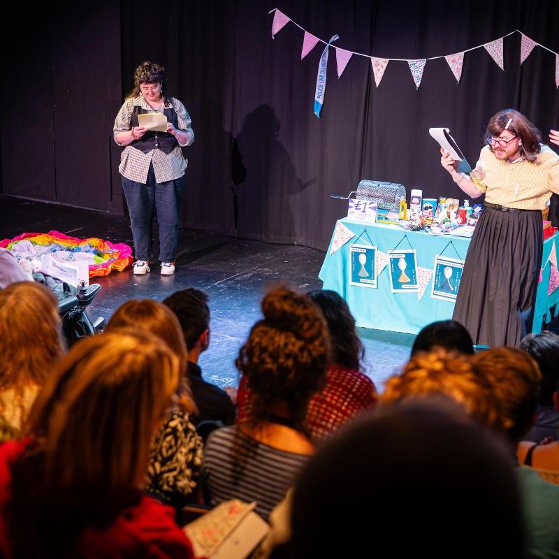 Two people are stood on stage in front of a tombola table and bunting reading from scripts, one has a brown bob and is in a yellow blouse and black skirt and the other has a brown mullet and vest. They are performing in front of a large audience