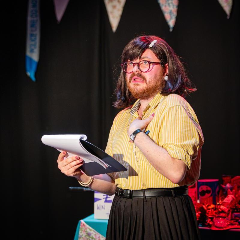 One person is stood in front of a tombola table lit red, they had a brown bob, are wearing a yellow blouse and are holding a clipboard, they look scared