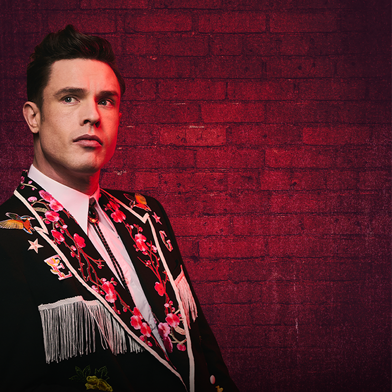 Ed Gamble stands in front of a red brick wall. He is dressed in a suit decorated with flowers and fringe.