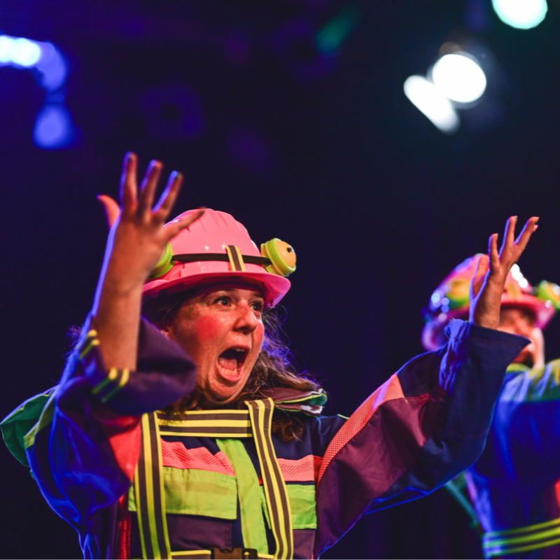 A woman is wearing a colourful costume and a hard hat that resembles a frog. She stands with her hands in the air with a shocked expression.