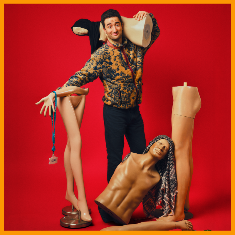 Hasan smiles surrounded by various pieces of mannequins, one wearing a black hijab and another wearing a keffiyeh, in front of a red background.