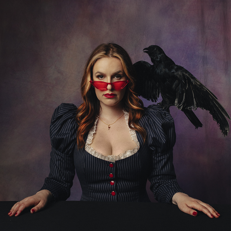 Katie Norris, a white woman with brown hair, stares into the camera. She is wearing a pair of red tinted sunglasses and a crow is perched on her shoulder.