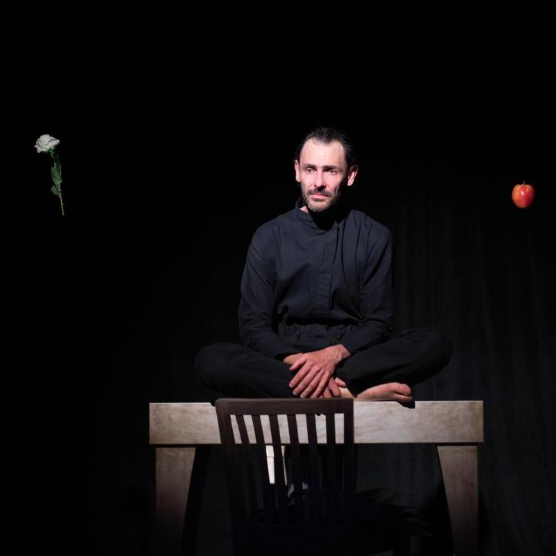 Photo of a man sat on a table, with a floating flower and apple next to his head