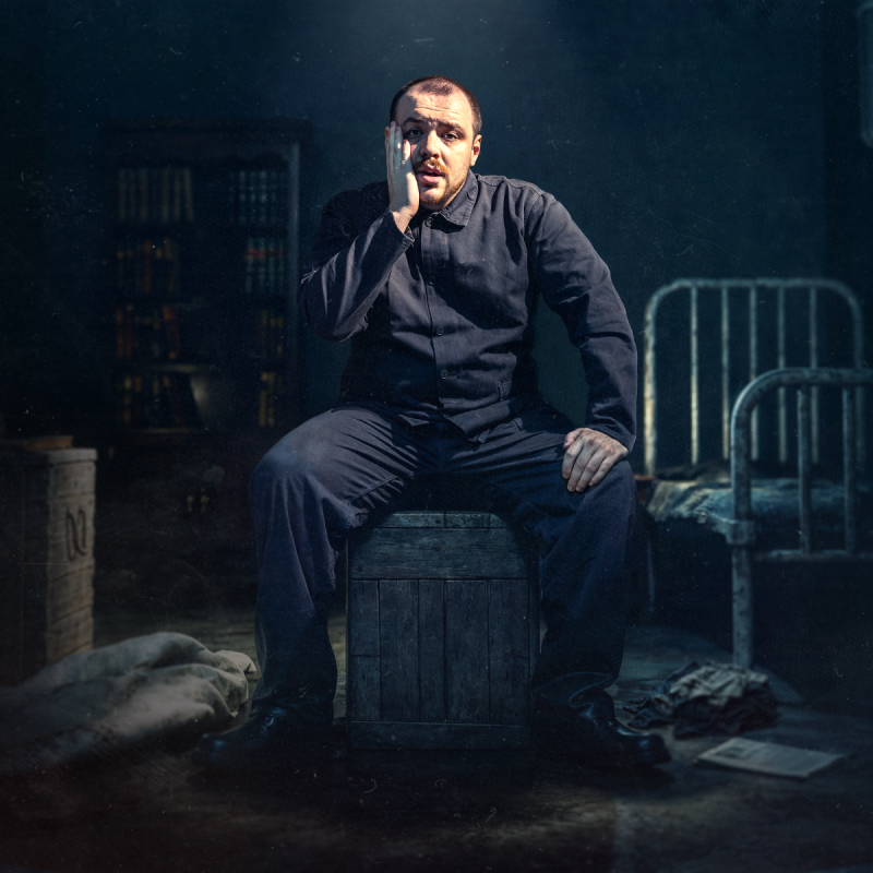 A person is in a dimly lit cell, sitting on a crate. They're wearing all-black and they look worried, holding a hand up to their bearded face. 
