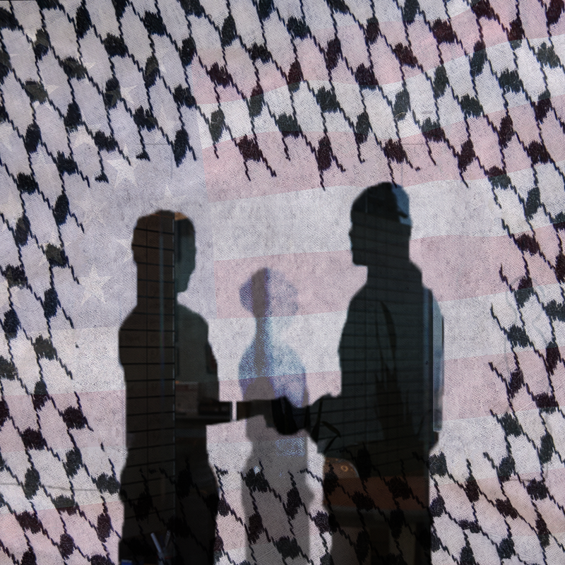 The shadows of two people holding hands. They can be seen in a cutout of fencing. In the background is a faded American flag.