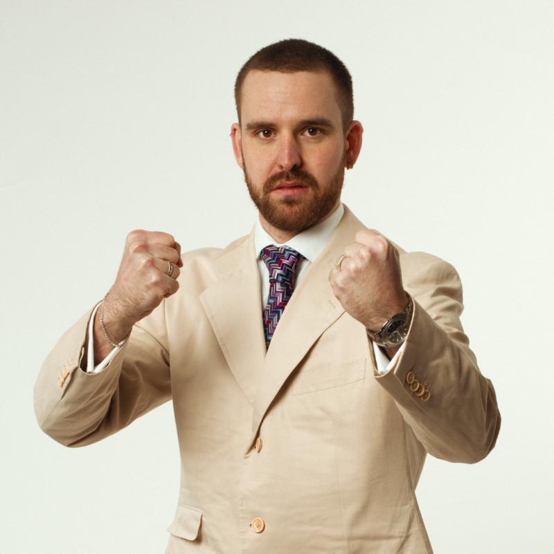Rob, in a pink suit jacket against a white background, holding up his fists combatively 