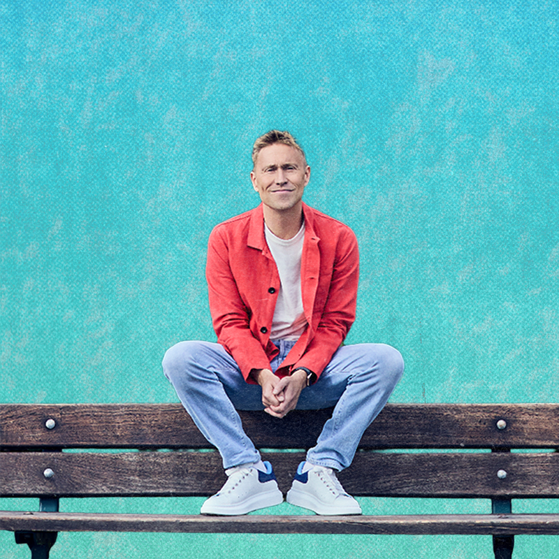 Russell Howard sits on the back of a bench in front of a blue backdrop. He has an amused expression.