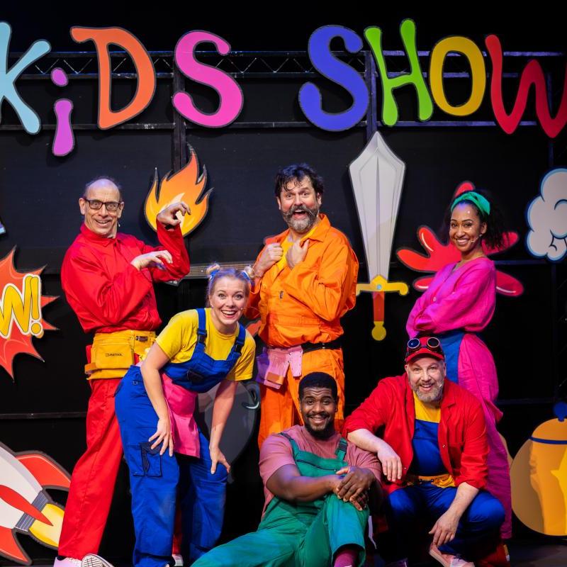 Image showing performers wearing bright colours in front of the 'Kids Show' sign, with cartoon props around them (e.g. a rocket and a sword)
