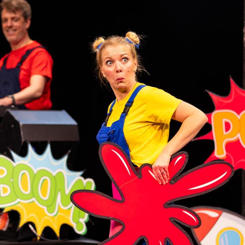 Photo of performer and musician behind, with signs that say 'BOOM!' and 'POW!'