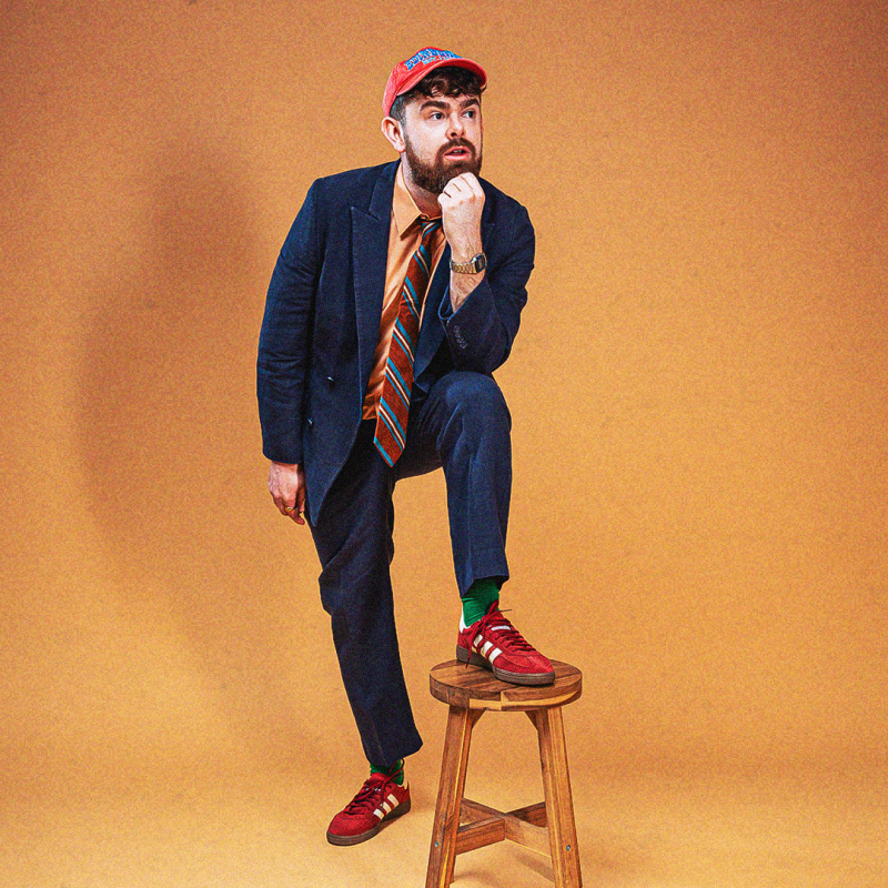 Tom is dressed in a navy suit, orange shirt, striped tie and red cap. He stands against an orange background with his foot perched on a wooden stool.