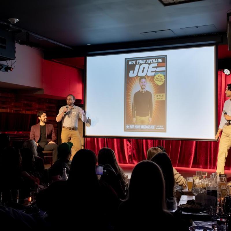 Three men standing on a stage. One of the men is holding a microphone and pitching his friend to the audience using a powerpoint presentation.