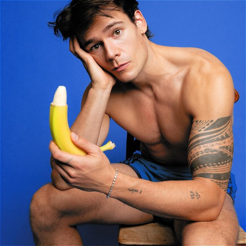 Mario Adrion sits shirtless in front of a blue background holding a partly peeled banana 