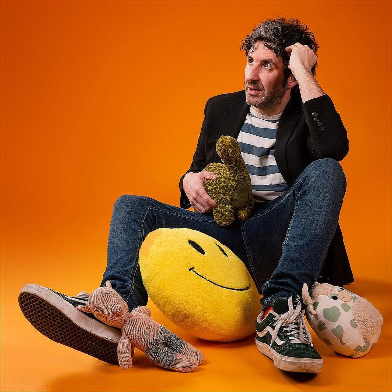 Mark Watson sitting on the floor holding children's toys and stuffed plushies.