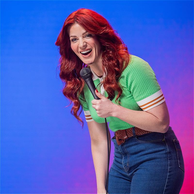 A woman dressed in a green shirt and blue jeans laughs into a microphone. Behind her is a bright pink and blue backdrop.