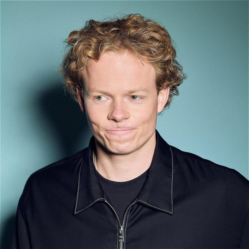 A curly haired man wearing a black zip up jacket gazes down with a mildly thoughtful expression. He is stood in front of a blue wall. 